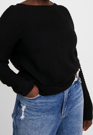 Personne portant un pull côtelé noir à manches longues et un jean bleu taille haute avec un patch usé, main dans la poche.