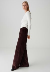 Maglietta bianca a maniche lunghe e pantaloni in corduroy bordeaux a gamba larga con vita elastica. La figura è in piedi di fronte a uno sfondo neutro.