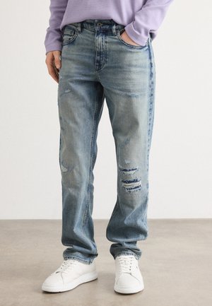 Light blue denim jeans with a relaxed fit, featuring distressed details and frayed patches, paired with white sneakers.