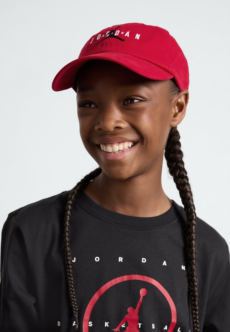 Căciulă de baseball roșie cu cuvântul "JORDAN" brodat în alb. Purtașul are împletituri lungi și este îmbrăcat cu un tricou negru cu un grafic de baschet roșu.