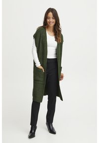 Hosszú, zöld, kötött mellény nyitott elülső résszel, bordázott felülettel és két elülső zsebbel, fehér hosszú ujjú inggel és fekete nadrággal viselve.