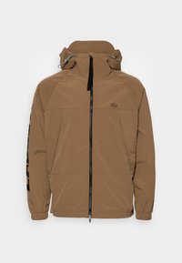 Brun vattentät jacka med hög krage, framdragkedja och justerbar huva. Har elastiska ärmslut och en logotyp på vänster ärm.