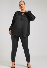 Svart, löst sittande blus med långa ärmar, rynkad halsringning och knytdetalj, kombinerad med figurnära svarta byxor och sandaler med remmar.