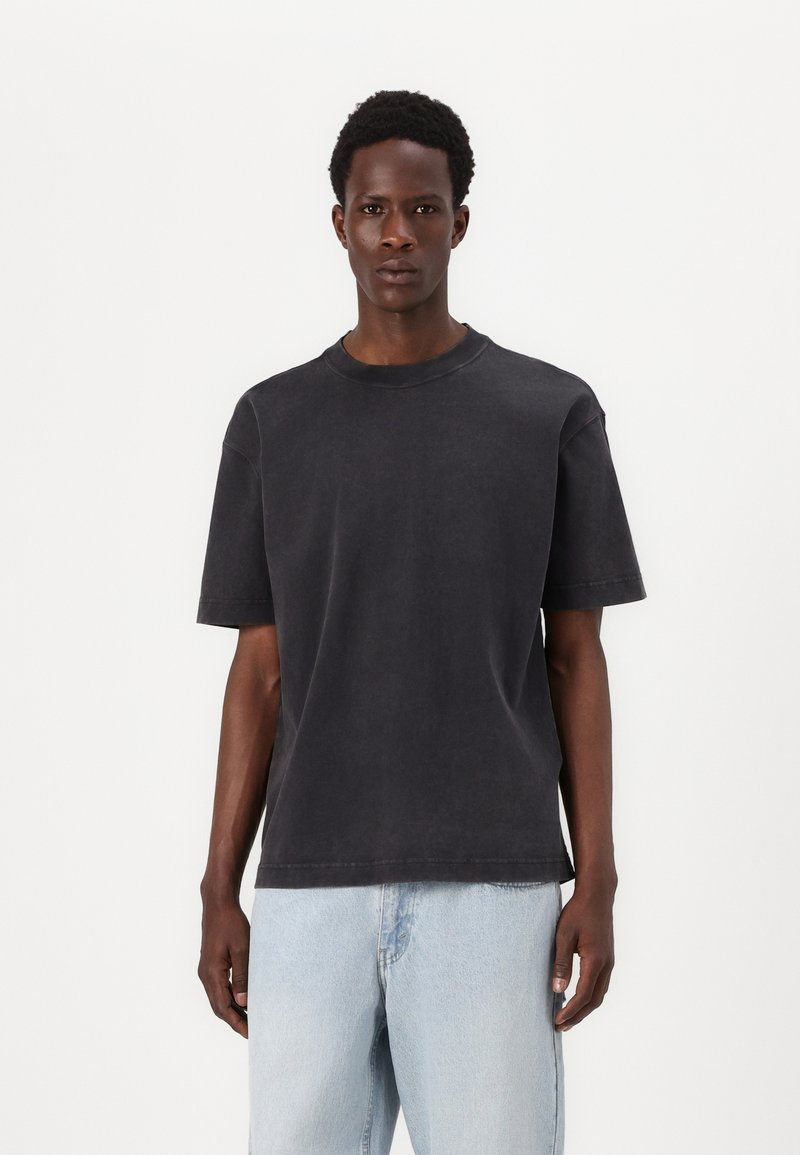 Tricou din bumbac de culoare gri închis, cu mâneci scurte, guler tip crew, croială lejeră și o textură ușor decolorată, asortat cu blugi de culoare albastru deschis.