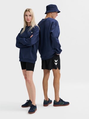 Blonde vrouw en man dragen marineblauwe sweaters en zwarte shorts; de man draagt een marineblauwe bucket hat, beiden in marineblauwe sneakers op een effen achtergrond.