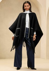 Poncho nero in lana con bordi frangiati, rifiniture bianche e due tasche frontali. Indossato sopra un dolcevita bianco e abbinato a jeans a gamba larga.
