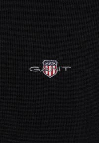 Tissu en maille noire avec un logo écusson brodé comportant des rayures rouges et blanches, des accents bleus, accompagné du texte "GANT".