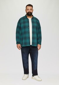 Mann steht, trägt ein türkisfarbenes karierte Hemd über einem weißen T-Shirt, dunkle Jeans und weiße Sneakers, vor einem schlichten hellen Hintergrund.