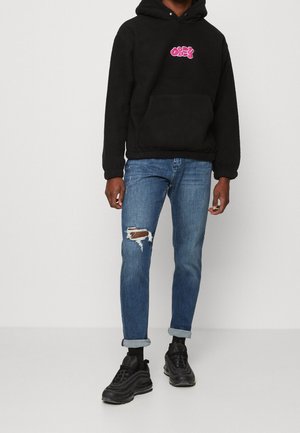 Henkilö, jolla on musta fleecehuppari punaisella logolla, kuluneet siniset käännetyt farkut ja mustat lenkkarit, seisomassa yksinkertaista taustaa vasten.