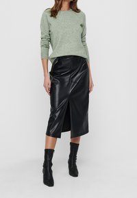 Hellgrüner Strickpullover mit langen Ärmeln, kombiniert mit einem schwarzen Kunstlederskirt in Midi-Länge, der einen vorderen Schlitz und strukturierte Nähte aufweist. Schwarze Knöchelstiefel.