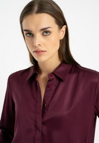 Bordowa satynowa bluzka z klasycznym kołnierzykiem, z zapięciem na guziki z przodu i długimi rękawami. Gładka faktura z lekkim połyskiem.