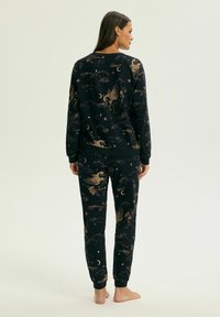 Kobieta ubrana w czarną bluzę z długim rękawem i spodnie w beżowe wzory z Pegazem, księżycem, gwiazdami i chmurami, stojąca boso, tyłem.