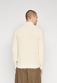 Suéter crema con un tejido texturizado, cuello alto y puños y dobladillo acanalados. Llevado con unos pantalones marrones de corte holgado.