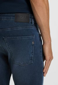 Mörkblå denimjeans i strukturerad tyg, med en svart läderlogotyp och en bakficka med präglad detalj.