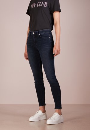 Persoon draagt donkere skinny jeans, zwart "PONY CLUB" t-shirt en witte sneakers, staand tegen een effen lichte achtergrond.