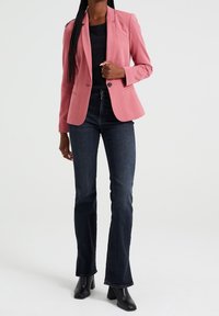 Růžový přizpůsobený blazer s jedním knoflíkem, spárovaný s tmavě modrými rozšířenými džíny. Blazer má klopové límce a dobře padnoucí střih.