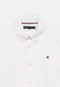 Λευκό πουκάμισο με κουμπιά φτιαγμένο από βαμβάκι, με απλωτό γιακά, περιλαμβάνοντας μπεζ κουμπιά και ένα μικρό λογότυπο σε μπλε, κόκκινο και λευκό στην περιοχή του στήθους.