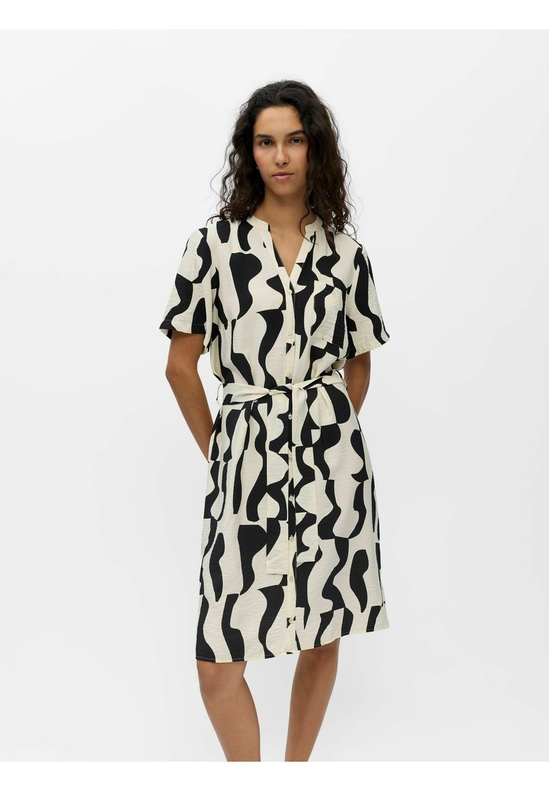 Femme portant une robe courte à manches courtes, longueur genou, avec des motifs abstraits noirs sur fond blanc, debout devant un fond blanc uni.
