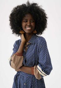 Blau gemustertes Kleid mit Rautenformen, langen Ärmeln mit braunen Akzenten, Knopfleiste vorne und strukturierten Manschetten. Einfache Gestaltung und weicher Stoff.