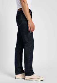 Jean en denim bleu foncé avec une coupe droite, texture subtile et couture claire. Portés avec des baskets blanc cassé avec des accents rouges.