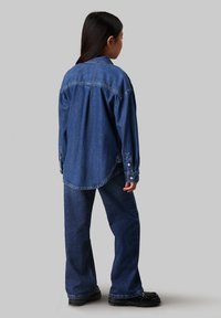Denim shirt and pants set in dark blue, featuring a relaxed fit, button details, and a curved hem on the shirt. Black footwear completes the look.