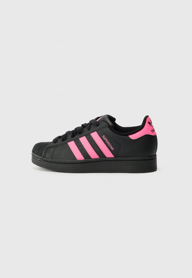 Fekete Adidas Superstar sportcipők, három rózsaszín csíkkal, textúrázott orral és fekete fűzővel, sima bőr felső résszel készültek.