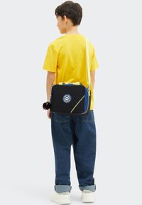 Sac bandoulière noir avec des accents bleus, fermeture éclair jaune et patch logo. Comprend un charm en fourrure attaché. Porté par un enfant en t-shirt jaune.