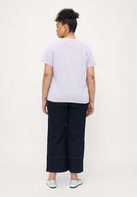 Camiseta de algodón de color púrpura claro con mangas cortas, combinada con pantalones de mezclilla oscuros de pierna ancha de talle alto y unos zapatos planos plateados, vista desde la parte de atrás.