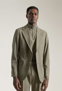 Traje verde oliva con chaqueta a medida y pantalones a juego. La chaqueta presenta solapas de muesca y cierre de un solo botón; la tela tiene una textura suave.