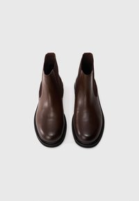 Botines de cuero marrón con paneles laterales elásticos, punteras redondeadas y suela plana negra, mostrando una textura suave y un diseño minimalista.