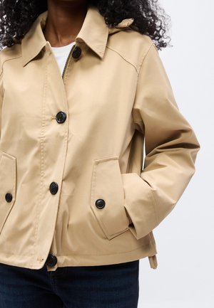 Kvinde iført en beige jakke med knapper og klaplommer, den ene hånd i lommen og mørkeblå jeans mod en ensfarvet lys baggrund.