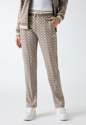 Donna che indossa pantaloni della tuta beige con fantasia e coulisse in vita, sneakers bianche e giacca coordinata con zip e righe sui polsini.