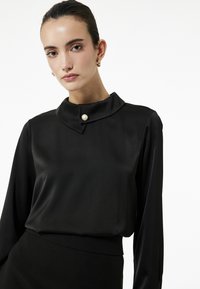 Schwarze Satinbluse mit hohem Kragen und einer Perlenknopf. Lange Ärmel und eine glatte Textur, entworfen für eine figurbetonte Passform.