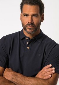 Mörkblå pikétröja med strukturerad tyg, tre träknappar vid kragen och en broderad hjortlogotyp på bröstet.