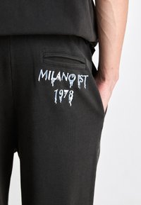 Sorte bukser med broderte hvite bokstaver "MILANO EST 1978" nær en baklomme, båret av en person med den ene hånden i frontlommen.