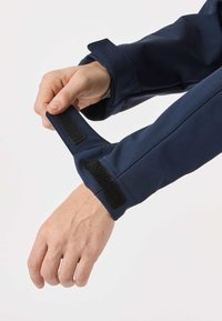 Person fastening black Velcro strap on navy blue jacket sleeve over light background.