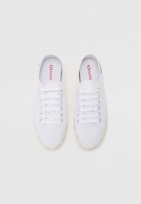 Par bijelih platnenih tenisica niskog profila s bijelim vezicama i ljubičastim "Superga" logotipom vidljivim unutar i na petnom dijelu, gornji pogled na bijeloj pozadini.