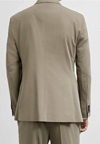 Uomo che indossa un giacca beige e pantaloni abbinati, visto di spalle su uno sfondo leggero e semplice.