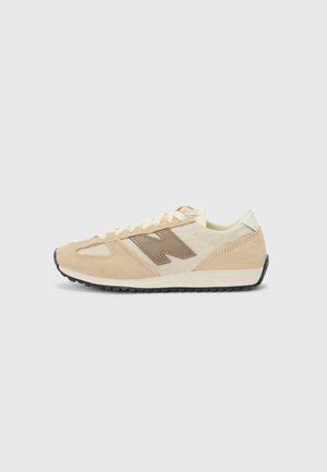 Low-profile beige and cream sneaker with suede overlays, lace-up closure, brown side branding, and black textured sole on a plain background.