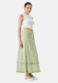 Hellgrüner Maxirock aus strukturiertem Stoff, mit gerüschten Stufen am Saum und einem Kordelzug in der Taille. Getragen mit einem weißen Cropped-Top und Schuhen.