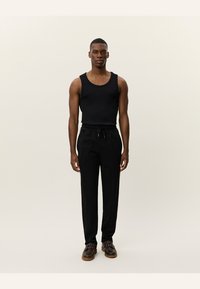 Schwarzes Tank-Top mit breiten Trägern, kombiniert mit schwarzen, schmal geschnittenen Hosen. Das Modell trägt dunkelbraune Schuhe mit strukturierter Sohle. Neutraler Hintergrund.
