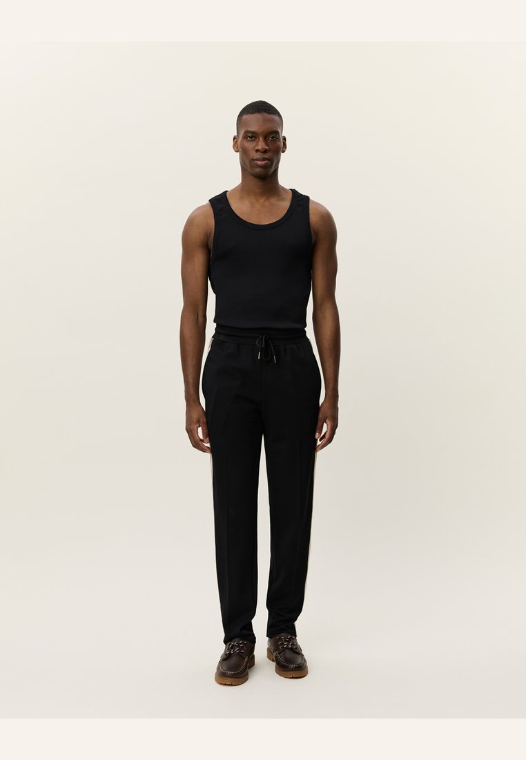 Schwarzes Tank-Top mit breiten Trägern, kombiniert mit schwarzen, schmal geschnittenen Hosen. Das Modell trägt dunkelbraune Schuhe mit strukturierter Sohle. Neutraler Hintergrund.