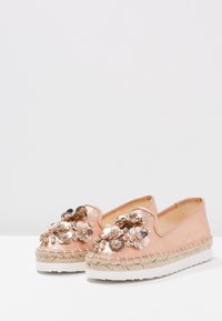 Espadrillas color oro rosa con decorazioni floreali, una suola in juta intrecciata e una parte superiore in tessuto liscio. Design slip-on con una base bianca piatta.