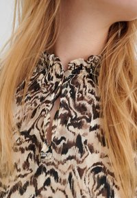 Blusa beige con un motivo astratto marrone, colletto arricciato, scollatura a chiave e un piccolo dettaglio con bottone decorativo. Texture liscia.