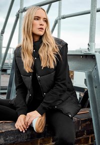 Gilet rembourré noir avec boutons-pression, porté par-dessus une chemise à manches longues noire, associé à des leggings noirs et des baskets blanches, assis sur un rebord.