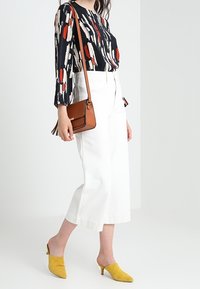 Flöda svart blus med abstrakt röd och krämfärgad mönster, kombinerad med vita vida byxor, gula klackar och en brun axelväska.