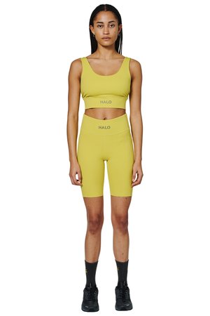 Kvinde, der står fremadvendt iført gul crop-top og matchende højttaljede cykelshorts med sorte sokker og sneakers på hvid baggrund.