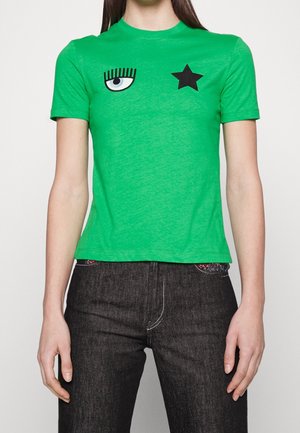 Grøn kortærmet skjorte med sort stjerne- og øjes grafik på brystet, båret med mørk denimjeans på en person, der står mod en hvid baggrund.