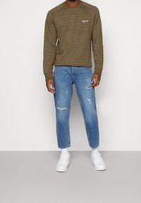 Olijfgroen textuur sweatshirt met lange mouwen en een witte scriptlogo, gecombineerd met lichtblauwe versleten spijkerbroeken en witte sneakers.