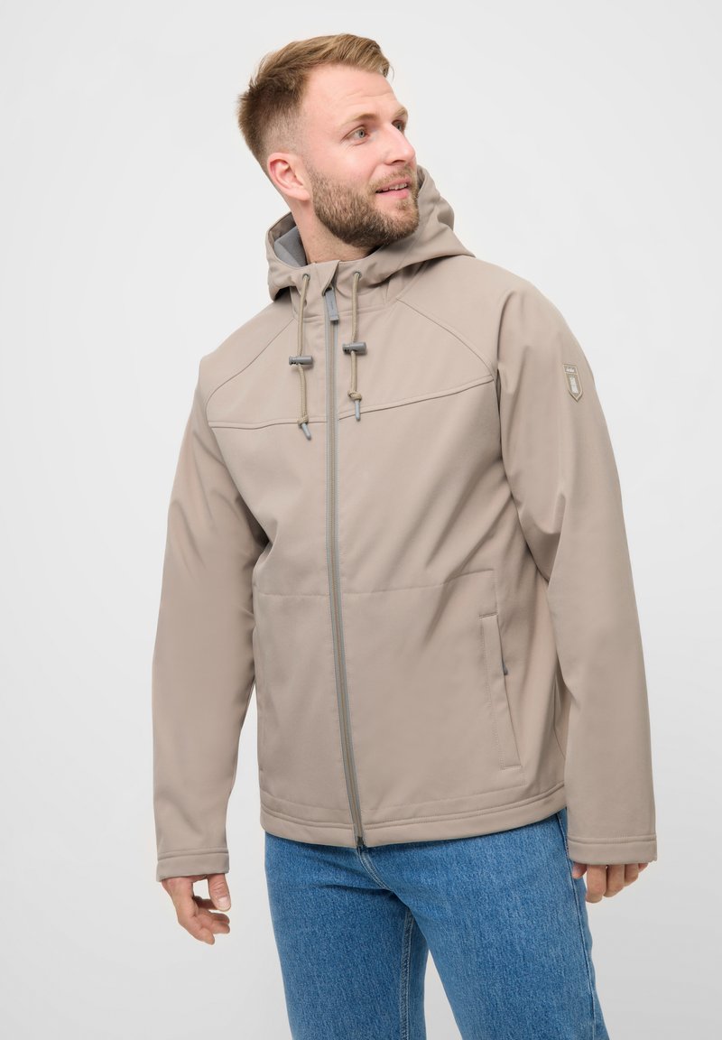 Mand med kort skæg iført beige hættejakke og blå jeans, der kigger mod venstre mod en ensfarvet baggrund.